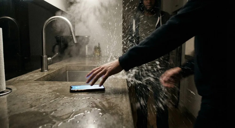 Water leaking from kitchen sink pipe while a hand reaches for smartphone to call emergency plumber