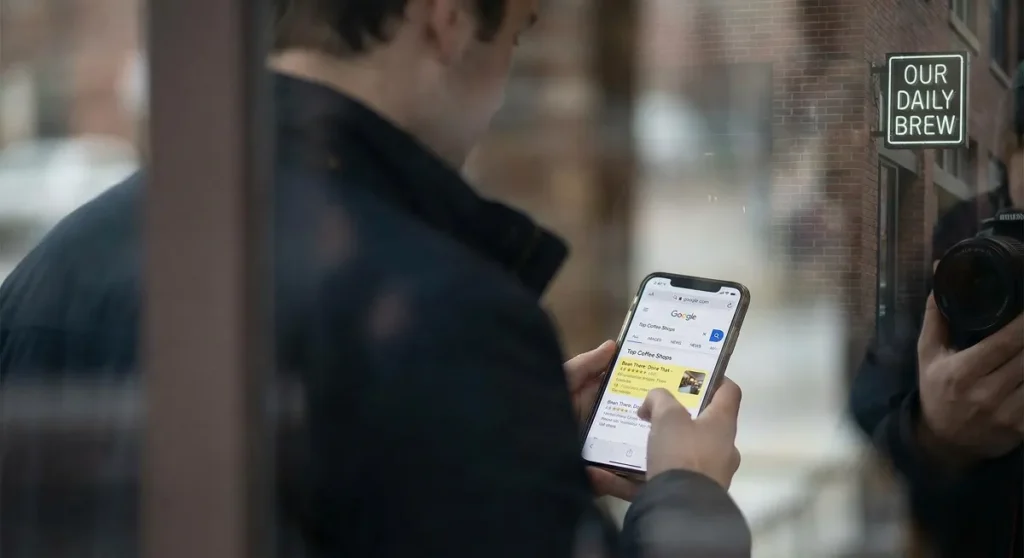 Business owner looking at phone showing competitor in Google search results with own shop sign blurred behind